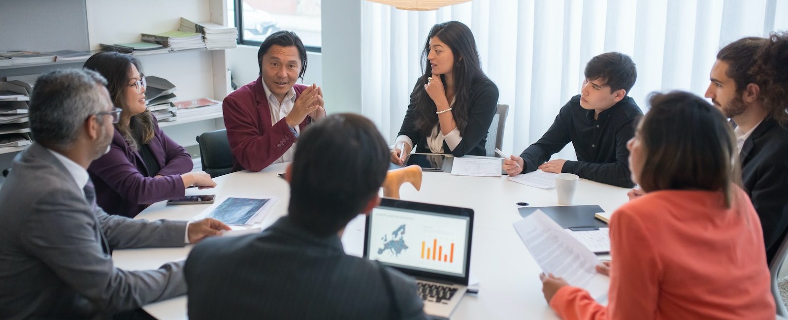 Group of professionals in a business meeting discussing strategies around a conference table with laptops and documents