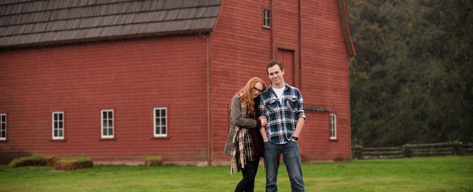 Couple standing in front of a rustic red barn on a grassy field with trees in the background