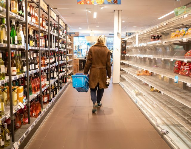 Shopper in a coat walks through supermarket aisle with wine bottles and empty shelves, carrying a basket and a bottle