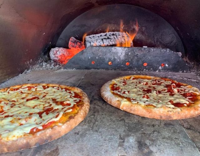 Two cheese pizzas baking in a traditional wood-fired oven with visible flames in the background