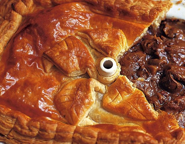 Golden steak and ale pie with rich filling, partially served in a white enamel dish for a classic British meal